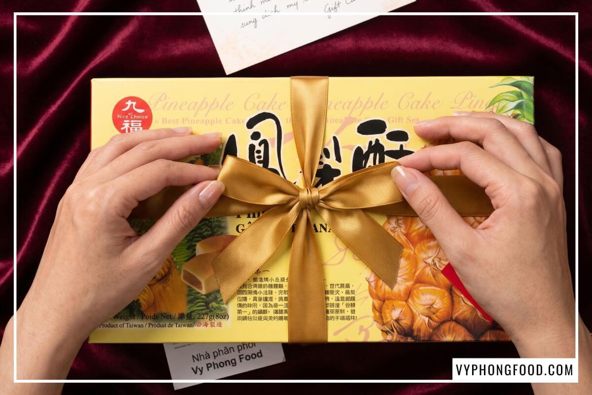 Hai bàn tay tỉ mẩn vuốt đuôi nơ ruy băng thắt chéo qua hộp bánh dứa Đài Loan Nice Choice nằm trên thảm nhung đỏ đô.