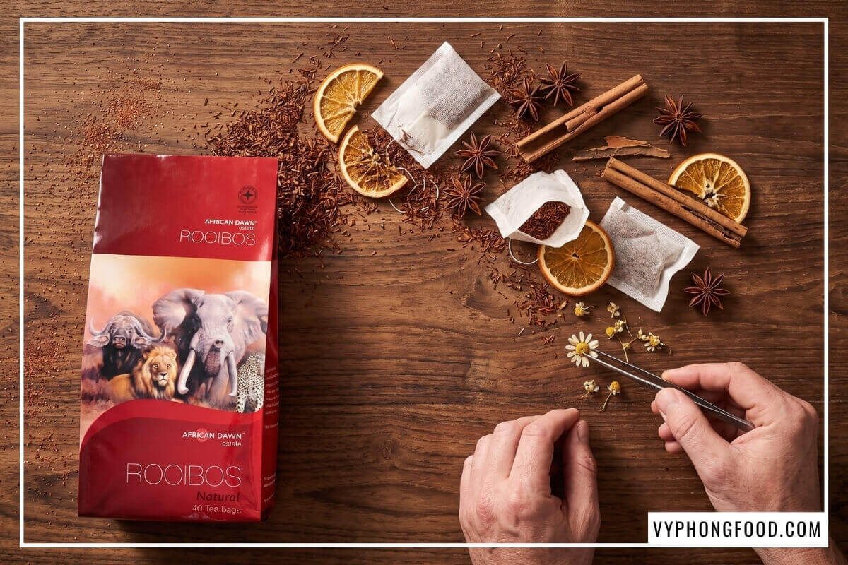 Mặt bàn gỗ bày lá trà đỏ khô, hoa hồi, thanh quế quanh gói trà rooibos túi lọc cao cấp chính hãng.