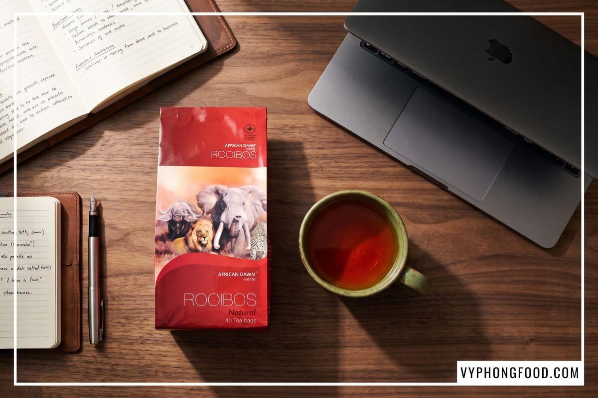 Flatlay tinh giản gói trà rooibos Nam Phi African Dawn đặt song song tách trà sứ xanh rêu và sổ tay trên bàn làm việc gỗ óc chó nét căng thớ gỗ tự nhiên.