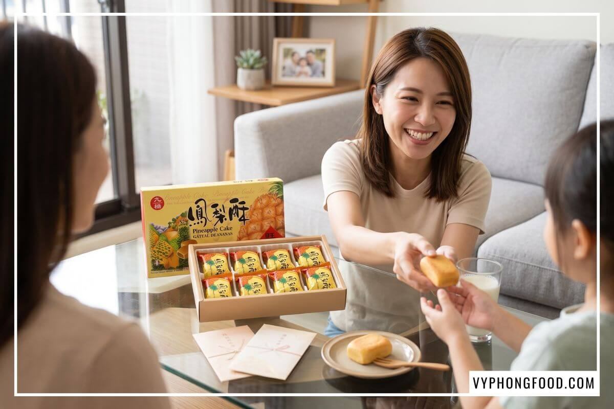 Người mẹ mỉm cười trao chiếc bánh dứa thơm bơ cho con gái nhỏ bên bàn trà phòng khách.