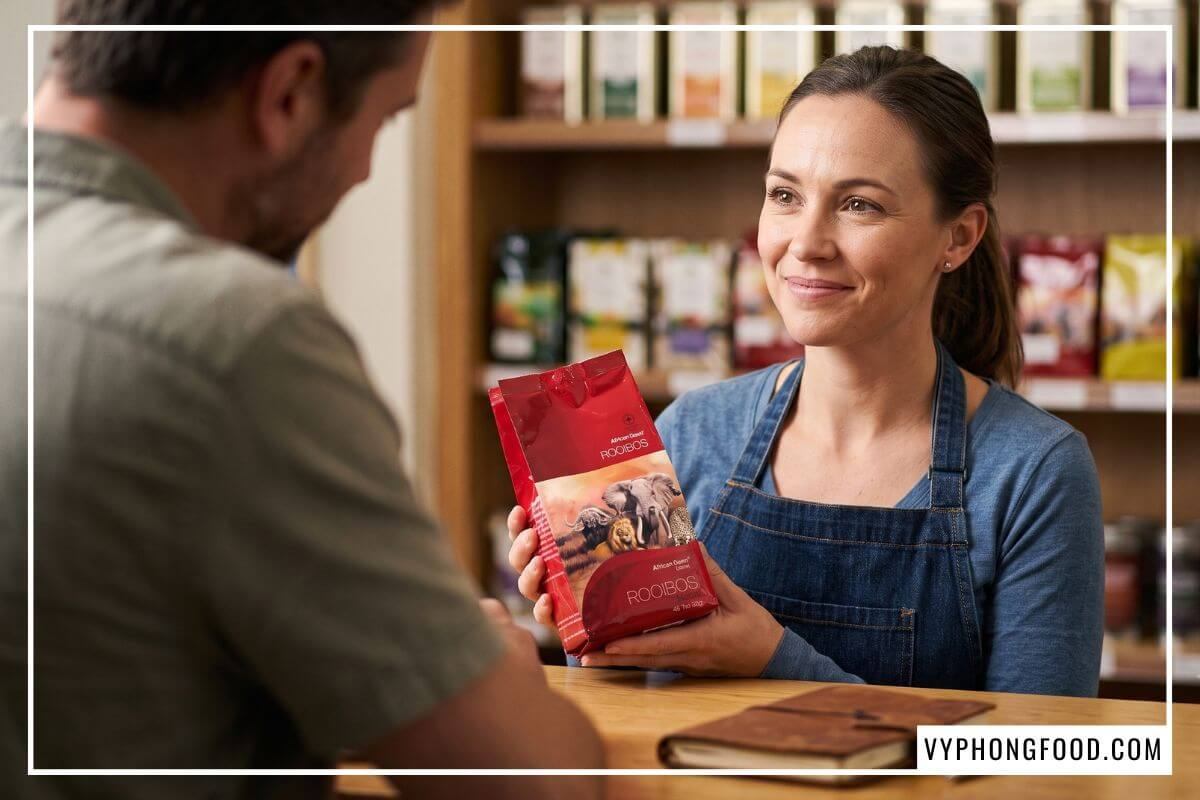 Khách hàng chăm chú nghe nữ nhân viên cầm hộp rooibos tea tư vấn trực tiếp tại cửa hàng bán trà thảo mộc