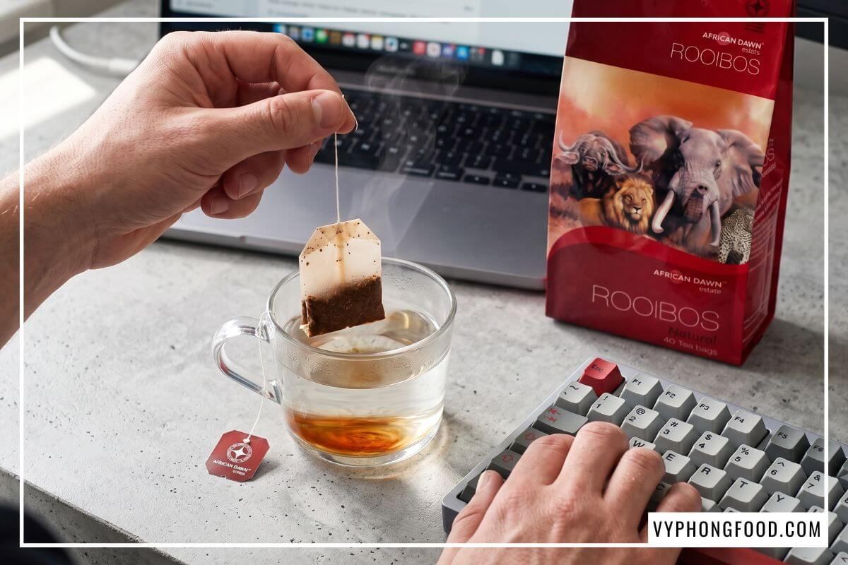 Một tay gõ phím, tay kia nhúng trà thảo mộc rooibos dạng túi lọc mua từ cửa hàng bán trà thảo mộc uy tín