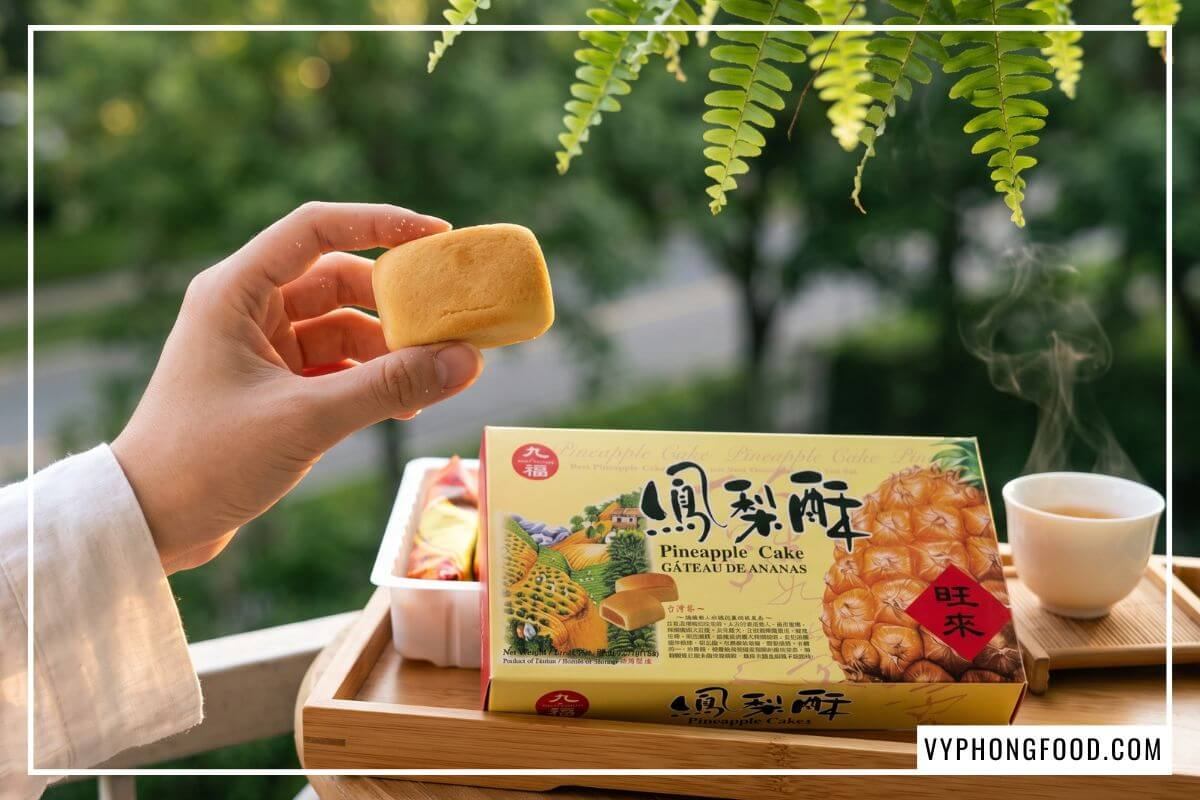 Tay nữ giới cầm chiếc bánh dứa Đài Loan chính hãng vàng ươm trước khay trà tre và hộp Nice Choice hé mở.