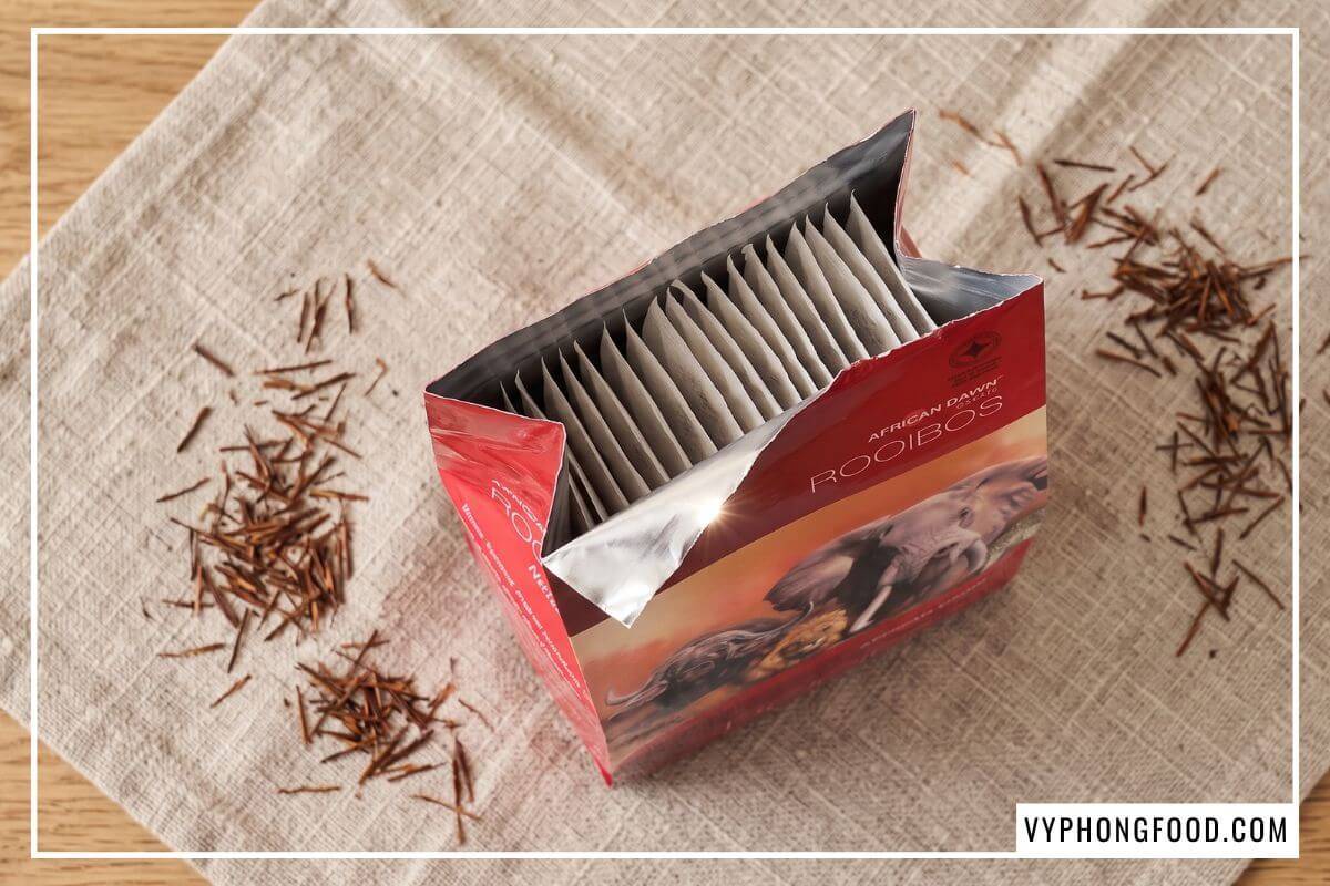 Hộp giấy đỏ mở nắp lộ túi lọc trà bên trong nằm trên vải linen thể hiện công dụng của trà thảo mộc Nam Phi nguyên bản.