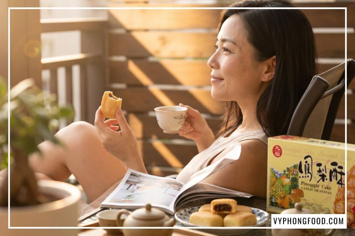 Người phụ nữ mỉm cười thư thái ngồi ở ban công đầy nắng cầm tách trà ấm và nếm nửa chiếc bánh dứa nướng.