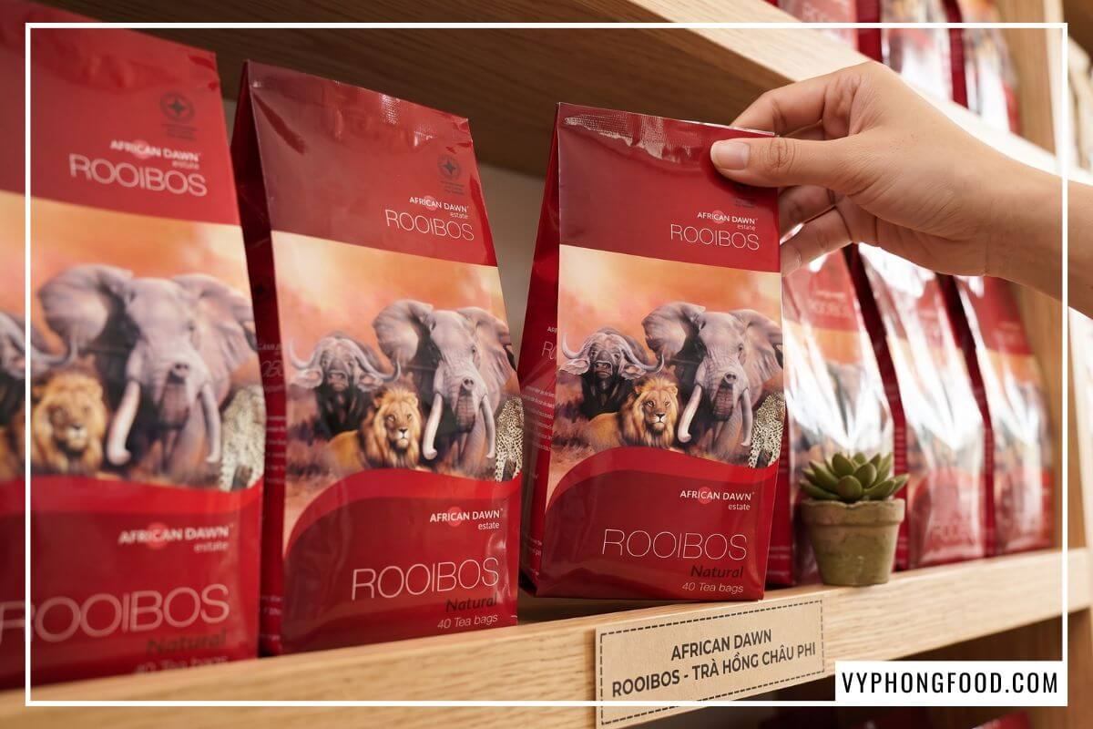 Khách hàng nhấc gói trà rooibos Nam Phi phẳng phiu trên kệ gỗ tại cửa hàng bán trà thảo mộc.