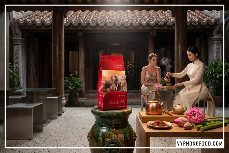 Nghệ nhân mặc áo dài cách tân pha trà bên đôn gốm cổ đặt dụng cụ đồng và gói trà túi lọc rooibos sỉ cho quán African Dawn nhập khẩu chính ngạch cạnh đĩa sợi trà khô, bông hoa sen hồng tươi và đốt tre xanh của Việt Nam.