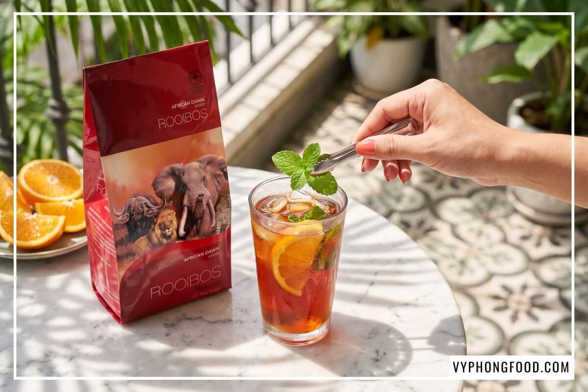 Bàn tay thả lá bạc hà vào ly cold brew cam vàng cạnh gói hồng trà rooibos Nam Phi giá tốt nhất African Dawn.