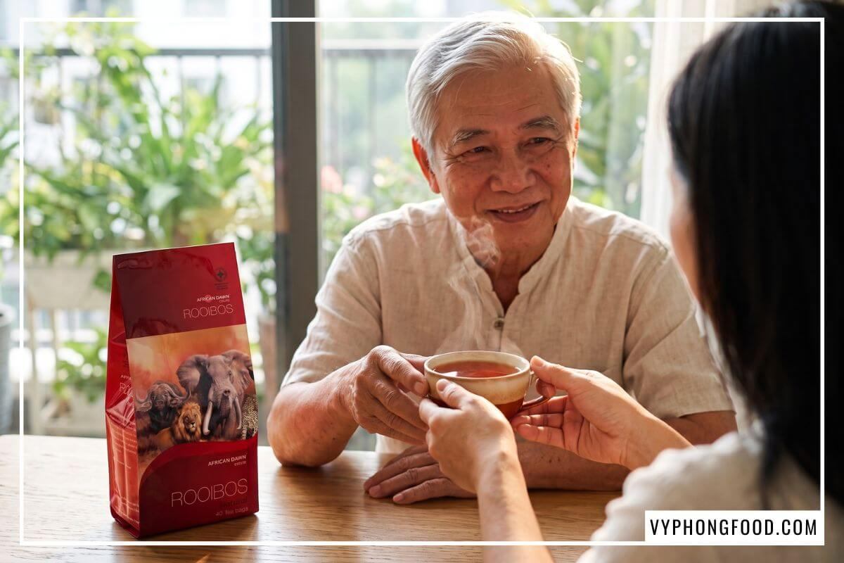 Người con gái trao tách trà rooibos đỏ hổ phách cho người cha lớn tuổi huyết áp cao bên gói trà African Dawn.