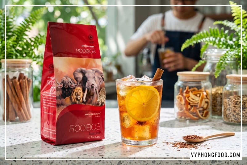 Gói trà rooibos giá sỉ African Dawn đặt cạnh ly cold brew cam quế mát lạnh trên quầy bar terrazzo TPHCM.