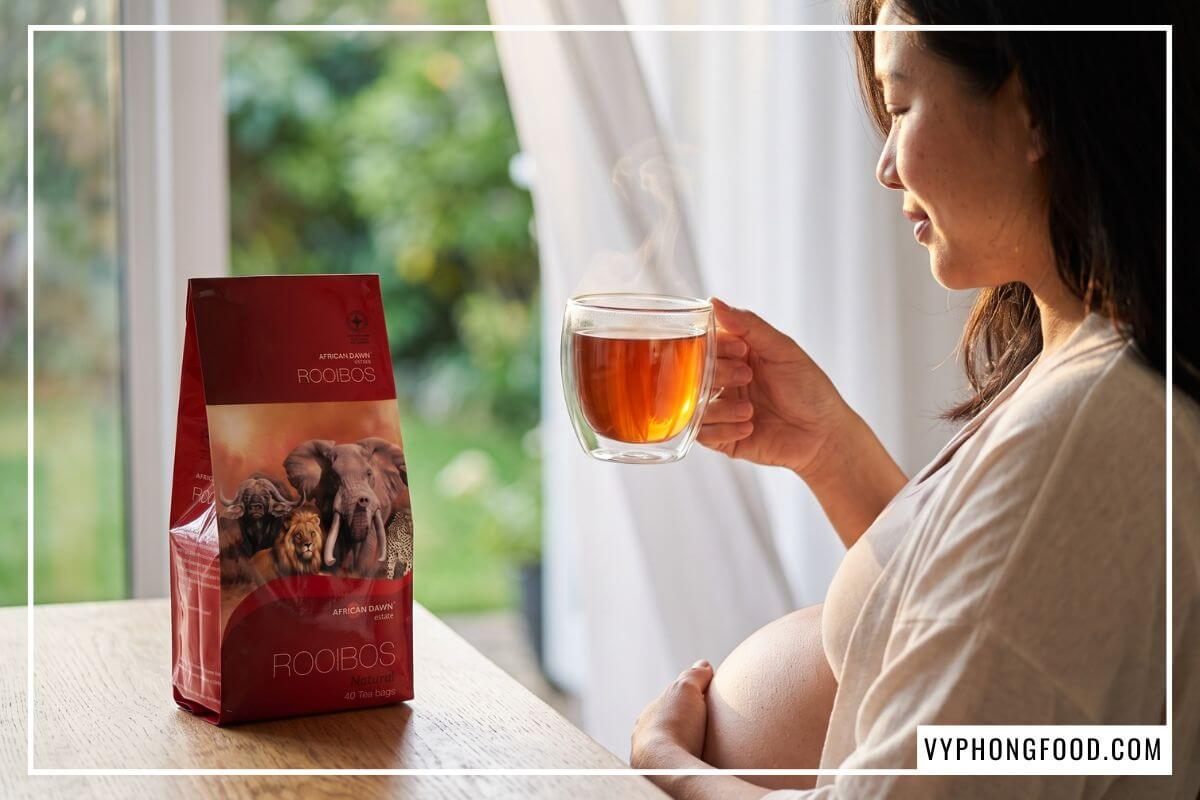 Mẹ bầu nâng tách trà rooibos túi lọc đỏ hổ phách bên gói trà African Dawn trên bàn gỗ ban công.