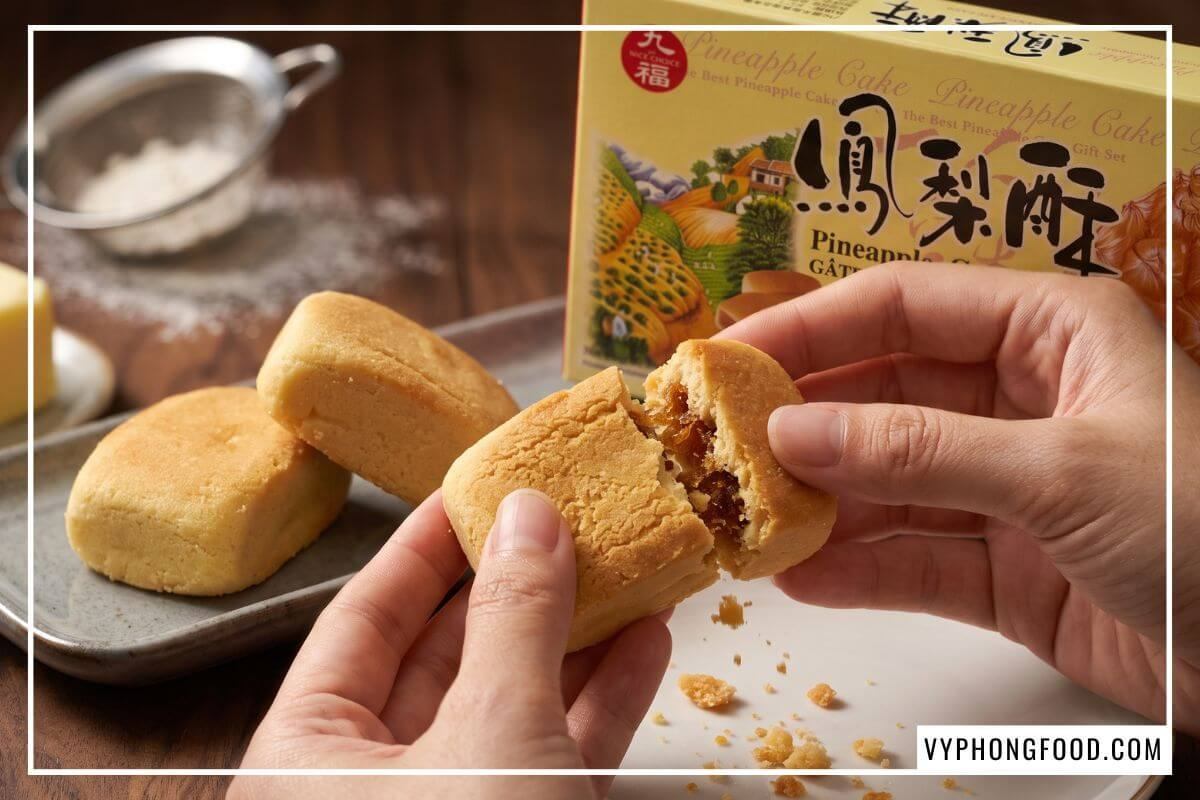 Tay bẻ đôi chiếc bánh nướng vàng ruộm làm rơi vụn bánh xốp giòn trên đĩa gốm cạnh hộp bánh dứa Nice Choice.