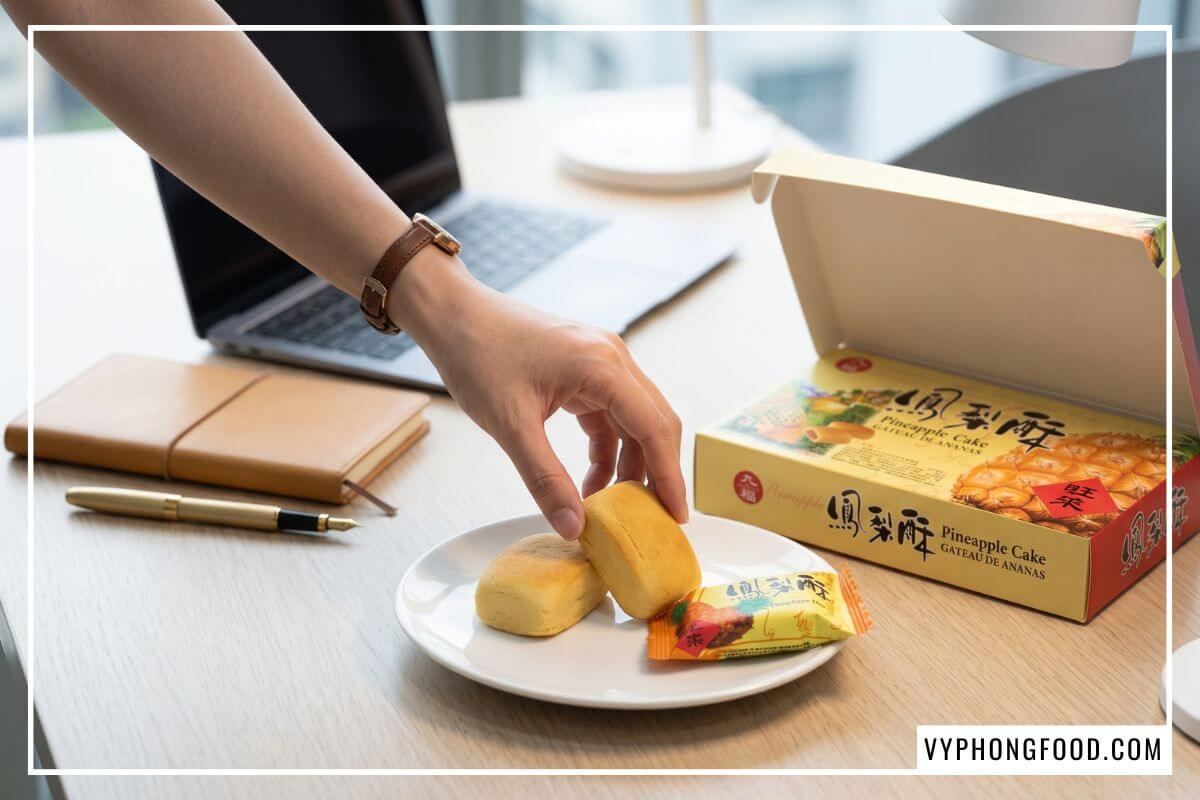 Tay nhân viên công sở lấy bánh dứa nướng trên đĩa sứ cạnh laptop mỏng và hộp bánh Nice Choice tại bàn làm việc.