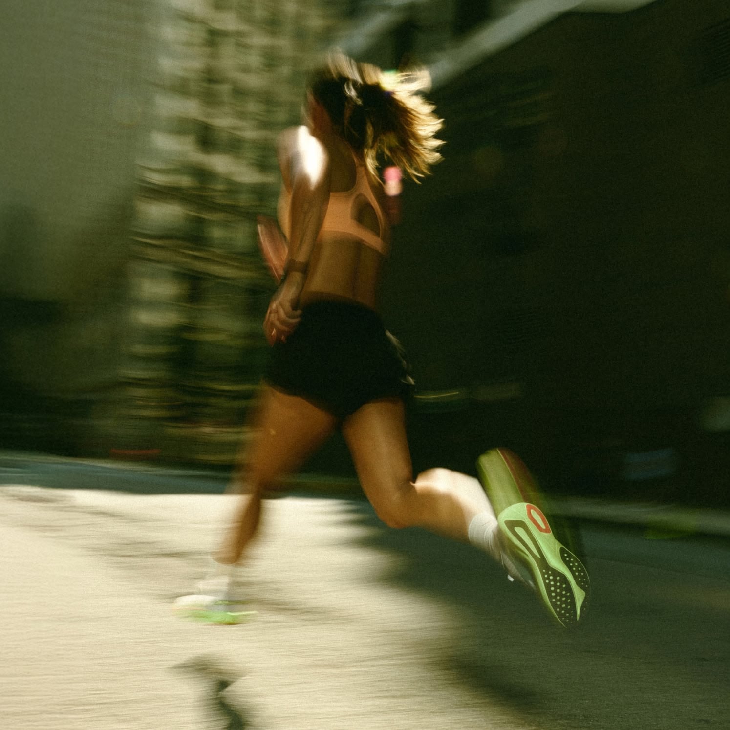Person running outdoors with motion blur, wearing a sports top and green shoes.