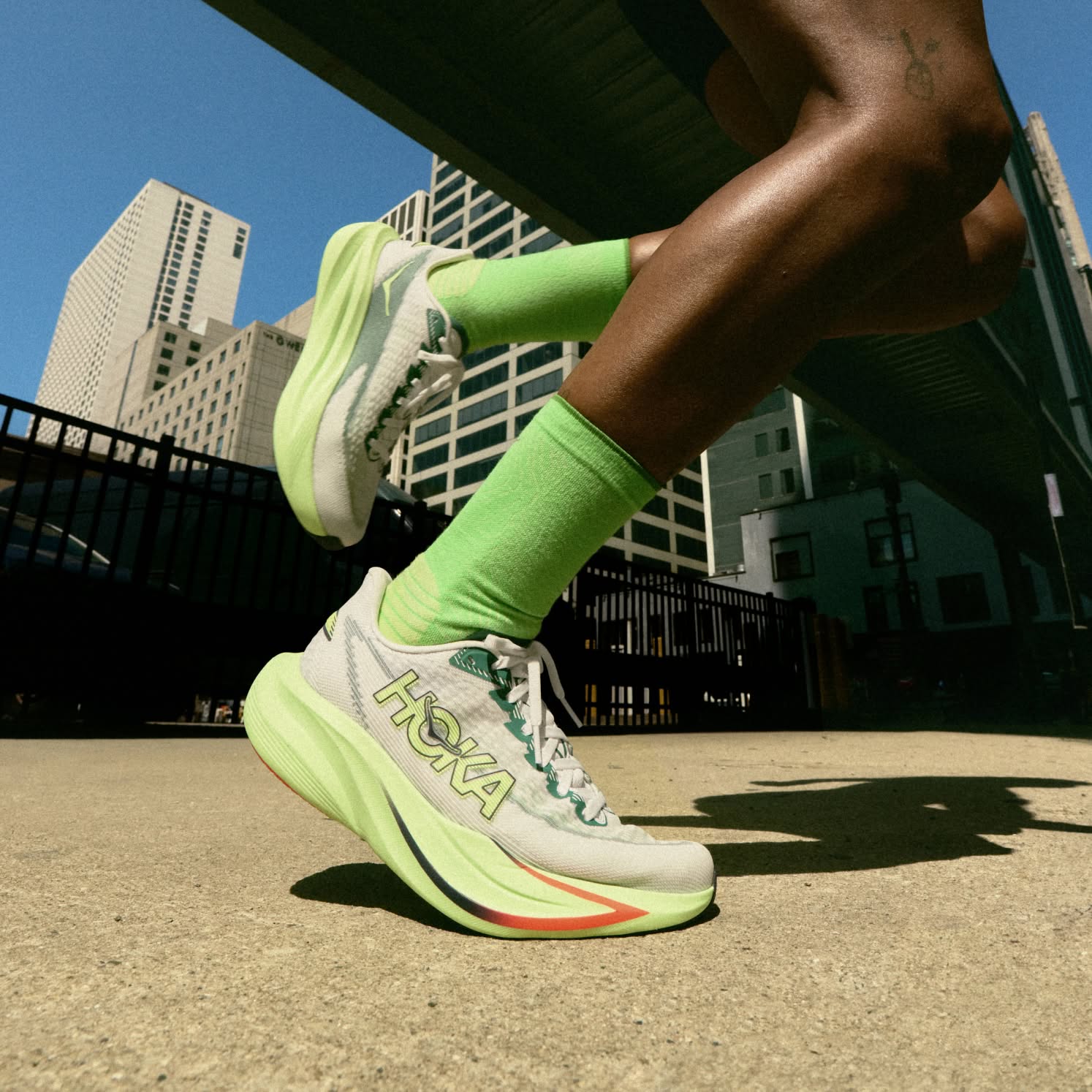 Runner in bright lime green Hoka running shoes and socks, mid-stride in a city.