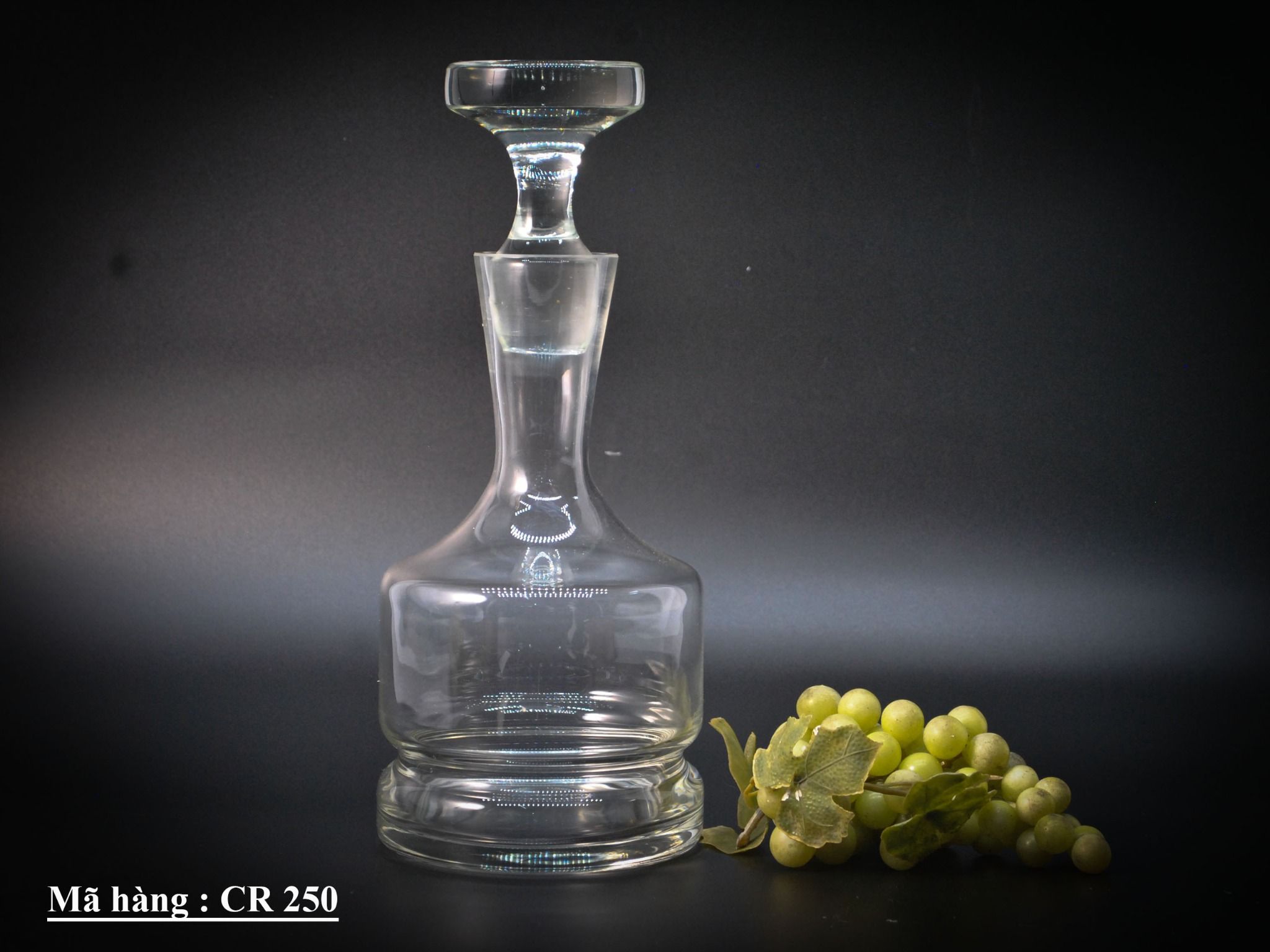 Một chiếc bình thủy tinh cổ điển dùng để đựng rượu, có thiết kế trong suốt với phần đế rộng và phần cổ hẹp, kèm theo nút đậy.
