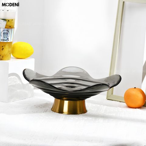 Tô trái cây thủy tinh lượn sóng đen khói đế vàng đặt trên bục trắng cạnh ly nước.