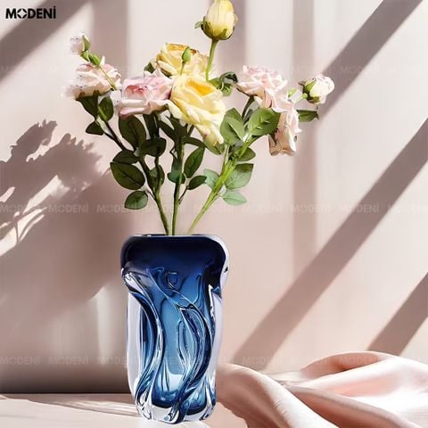 Góc nhìn chính diện bình hoa thủy tinh kiểu dáng sóng nước màu xanh dương cắm hoa hồng đặt trên dải lụa mềm.