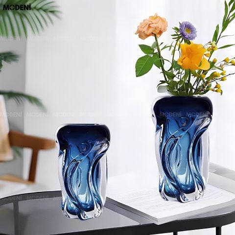 Góc nhìn chính diện bình hoa thủy tinh kiểu dáng sóng nước màu xanh dương cắm hoa hồng đặt trên dải lụa mềm.