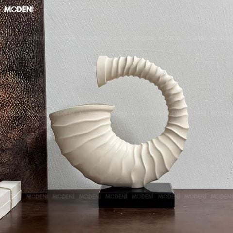 Trưng bày trên bàn gỗ tối màu, tượng trang trí Voluta màu be nổi bật với đường xoắn vỏ ốc nghệ thuật bên bức tranh kim loại.