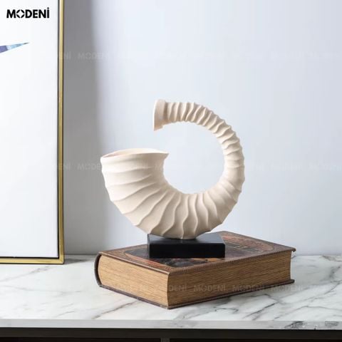 Trưng bày trên bàn gỗ tối màu, tượng trang trí Voluta màu be nổi bật với đường xoắn vỏ ốc nghệ thuật bên bức tranh kim loại.