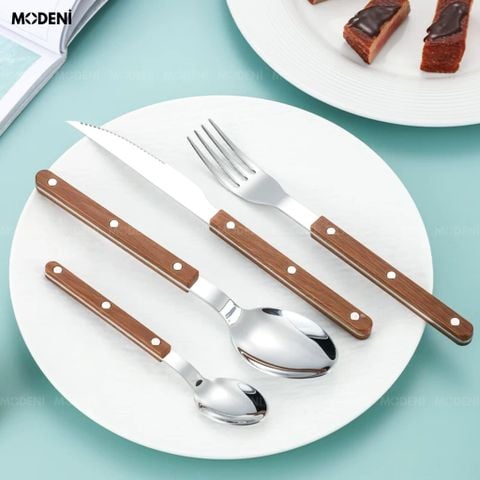 Bộ dao muỗng nĩa Bresso Inox trên đĩa sứ trắng làm nổi bật sự đối lập giữa đầu bạc sáng bóng và cán gỗ nâu mộc mạc.