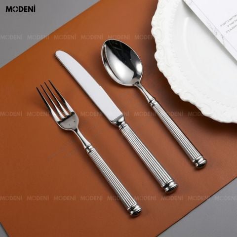 Bộ dao muỗng nĩa Silveria Inox 304 ánh bạc đặt chéo nghệ thuật trên đĩa sứ trắng họa tiết cổ điển với dải ruy băng đen.