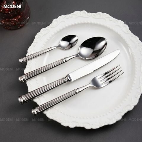 Bộ dao muỗng nĩa Silveria Inox 304 ánh bạc đặt chéo nghệ thuật trên đĩa sứ trắng họa tiết cổ điển với dải ruy băng đen.