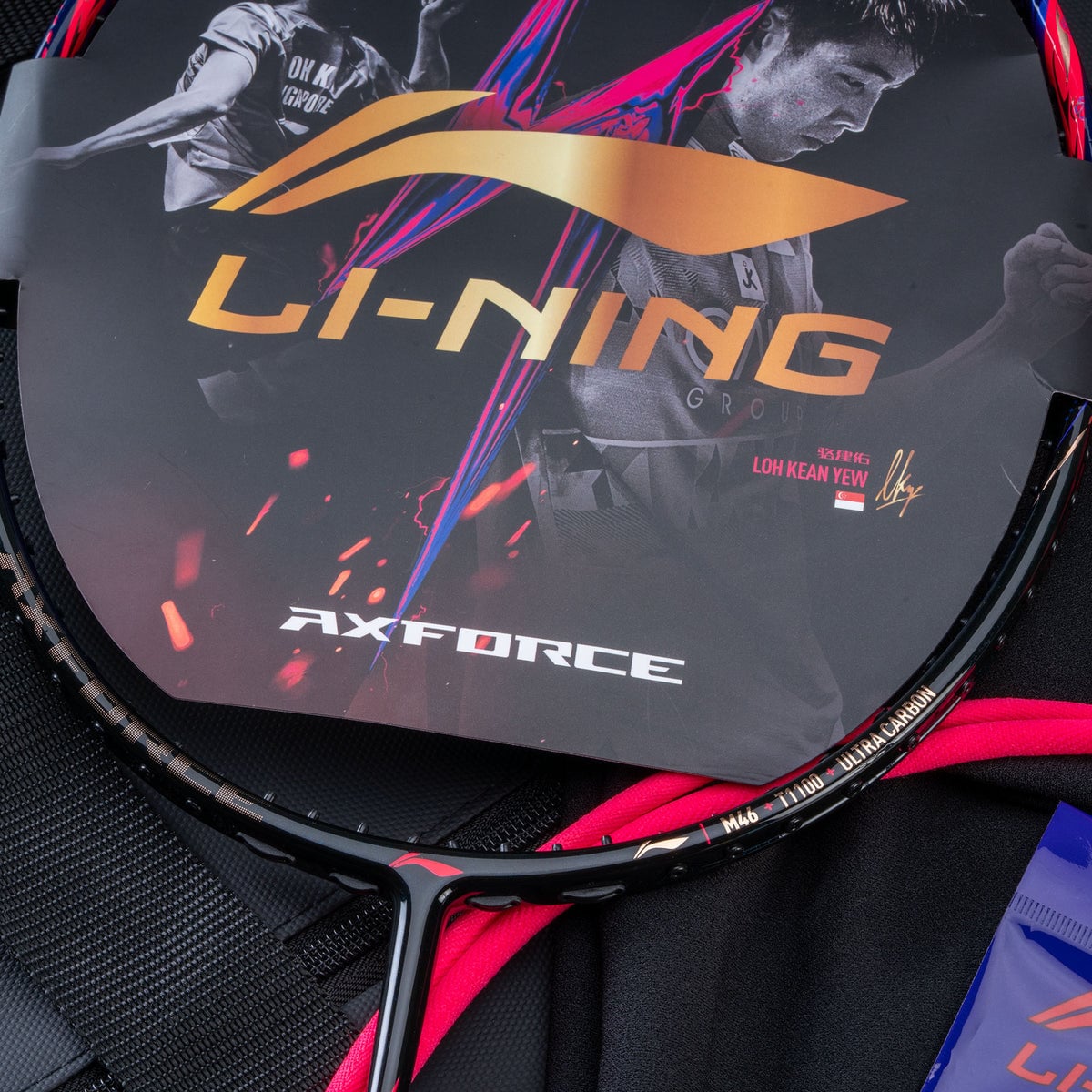 Cận cảnh khung vợt Lining Axforce 90 LKY đặt cạnh bao vợt in logo LI-NING AXFORCE và chữ ký Loh Kean Yew kèm cờ Singapore, cạnh khung in chữ M46 T1100 Ultra Carbon