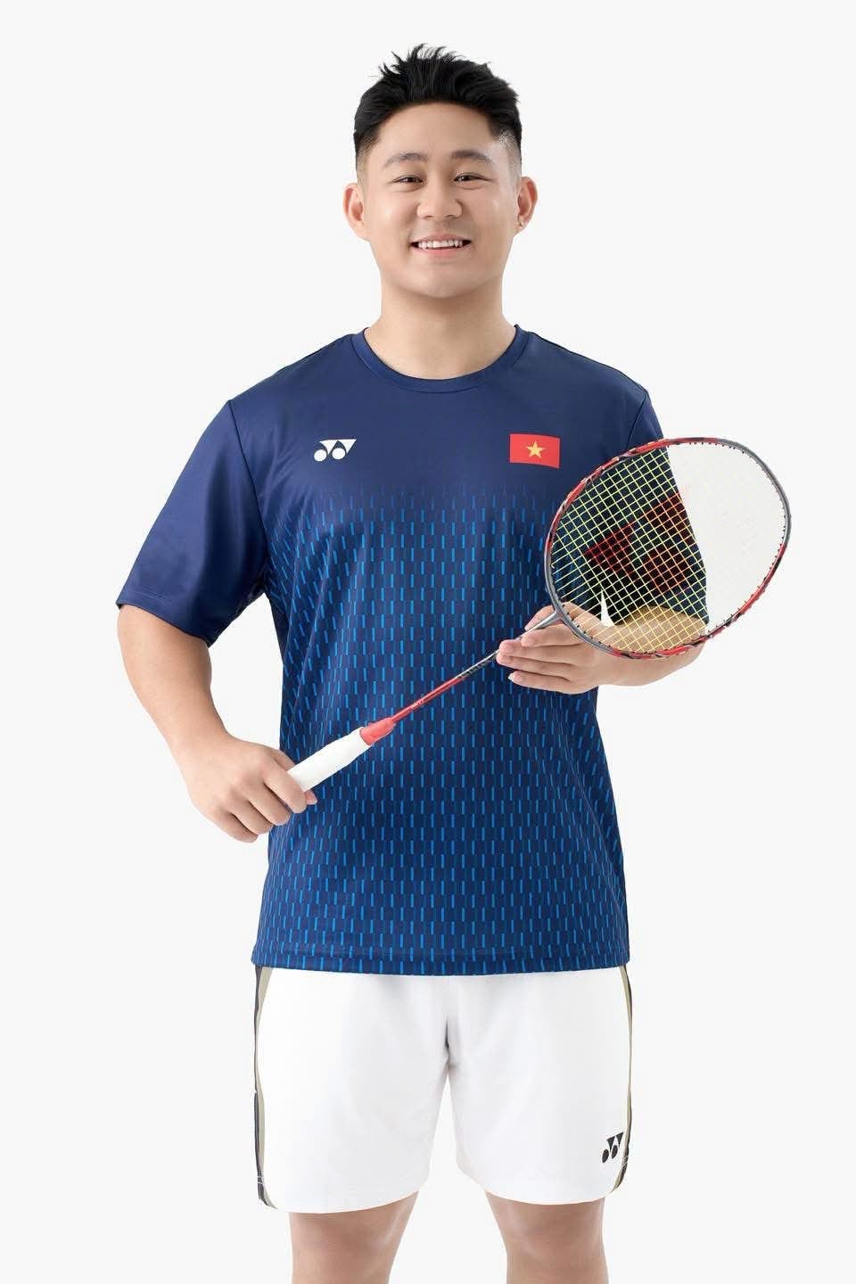Người mẫu nam mặc áo cầu lông Yonex Vietnam Open TRMYOB26147Z bản Dark Navy màu xanh navy đậm, logo YONEX trắng ở ngực trái, cờ đỏ sao vàng Việt Nam ở ngực phải, họa tiết vảy chìm cùng tông ở phần thân dưới, mặc cùng quần short trắng