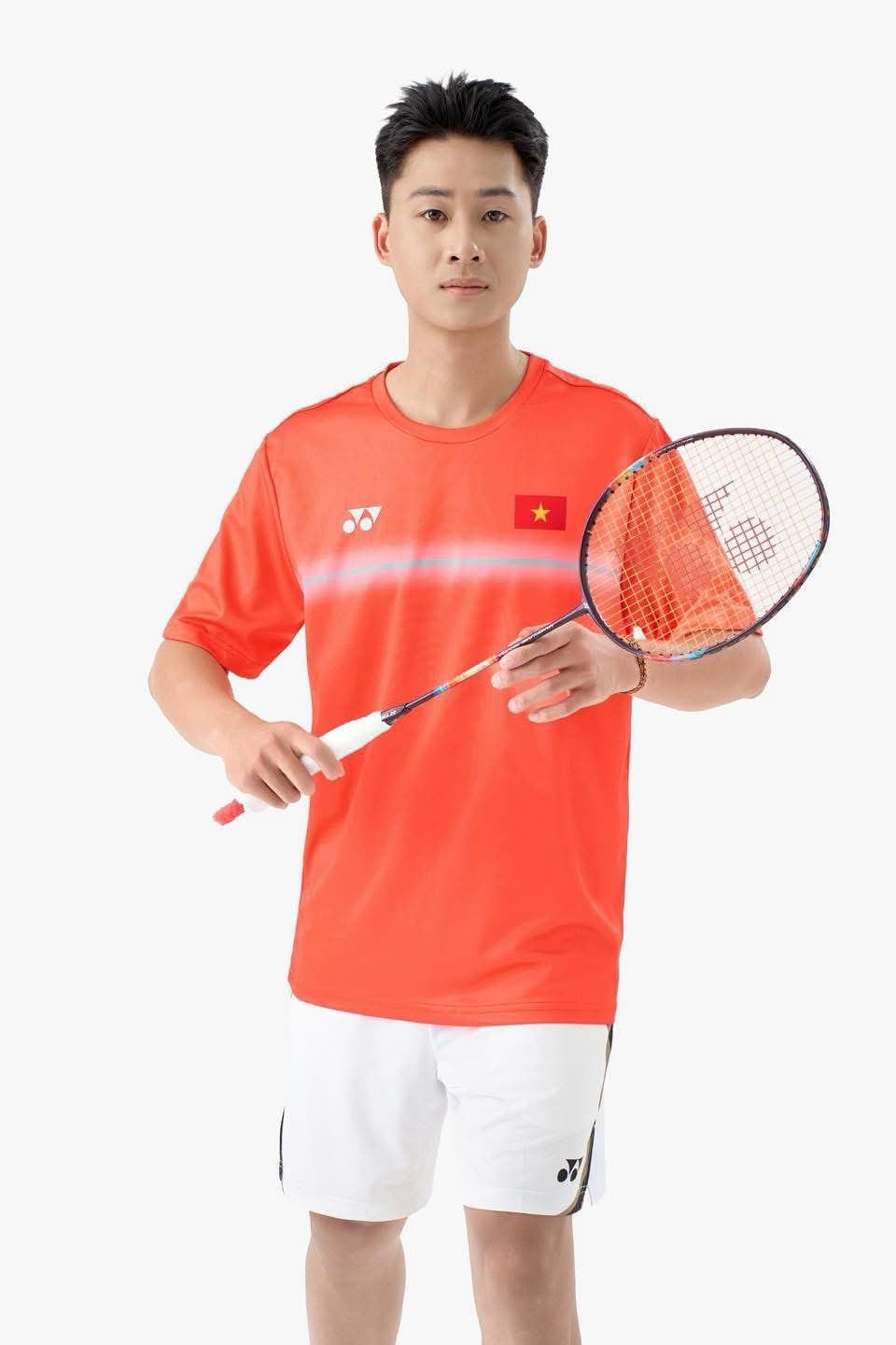Người mẫu nam mặc áo cầu lông Yonex Vietnam Open TRMYOB26147Z bản Cyber Orange màu đỏ cam, logo YONEX trắng ở ngực trái, cờ đỏ sao vàng Việt Nam ở ngực phải, dải chuyển sắc trắng xanh nhạt chạy ngang ngực, mặc cùng quần short trắng